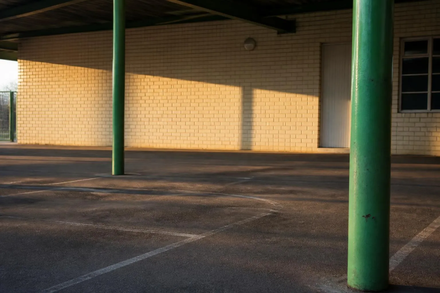 Un préau d'école élémentaire vide, ses poteaux métalliques peints et le sol de bitume marqué au blanc