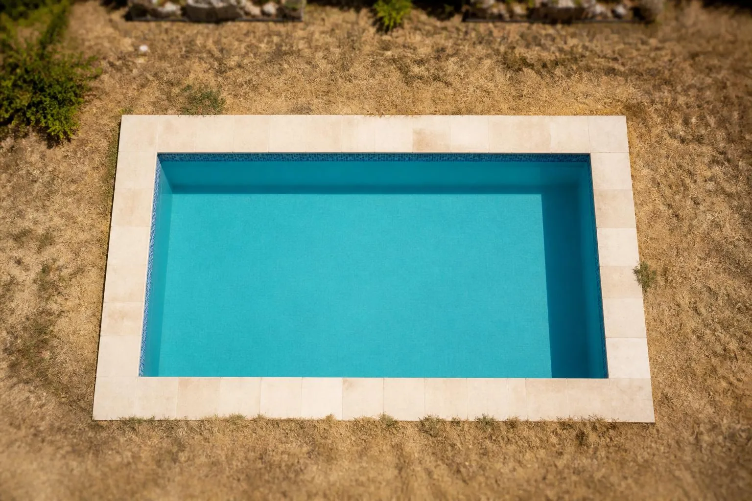 Un bassin enterré rectangulaire vu du dessus, margelles en pierre claire et eau bleue immobile, entouré d’une pelouse sèche.